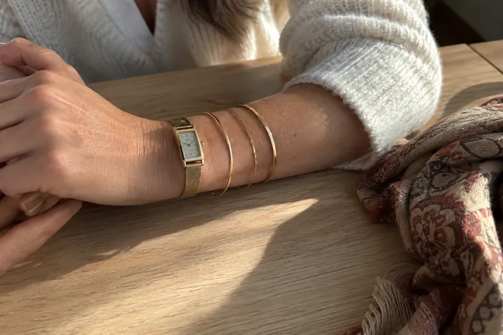 A woman's wrist wears a rectangular gold watch and three thin gold bangles, resting on a pale wooden table beside a patterned scarf.